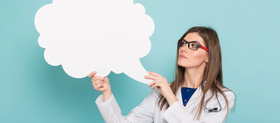 Portrait of female doctor in white coat and glasses wears stethoscope thinks about treatment holds blank paper thinking speech bubble isolated on blue background wondering medical issues concept.